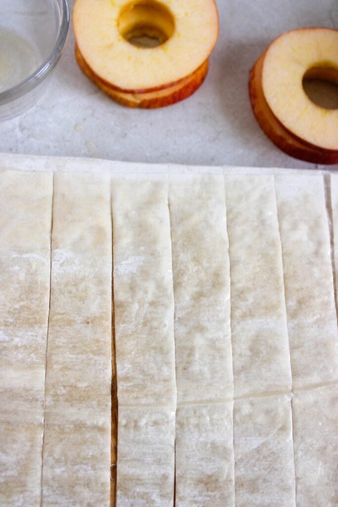 Slices of puff pastry dough arranged in rows on a surface next to apple slices with cores removed, ready to be transformed into delicious cinnamon apple strudel slices.