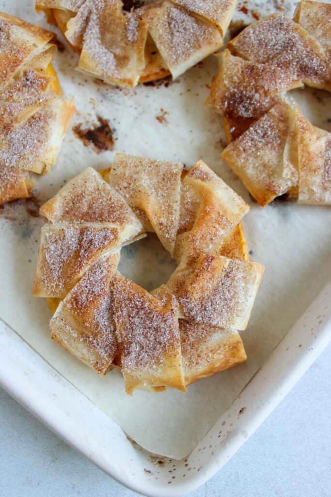Crispy, sugar-dusted pastry rings, reminiscent of cinnamon apple strudel slices, are arranged on a parchment-lined baking tray.