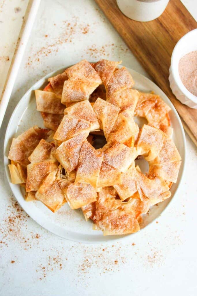 A plate of cinnamon apple strudel slices dusted with cinnamon sugar sits on a white surface, next to a wooden board with a bowl of additional cinnamon sugar.