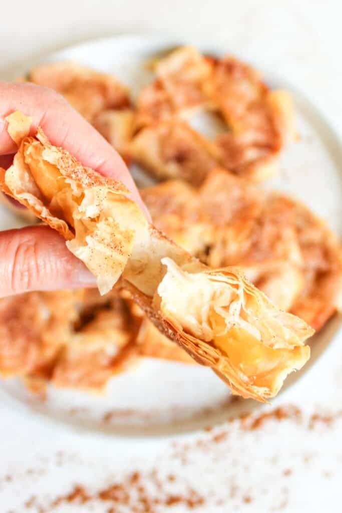 A hand holds a piece of flaky pastry filled with apple filling, evoking the essence of Cinnamon Apple Strudel Slices, with more pastries on a white plate in the background.