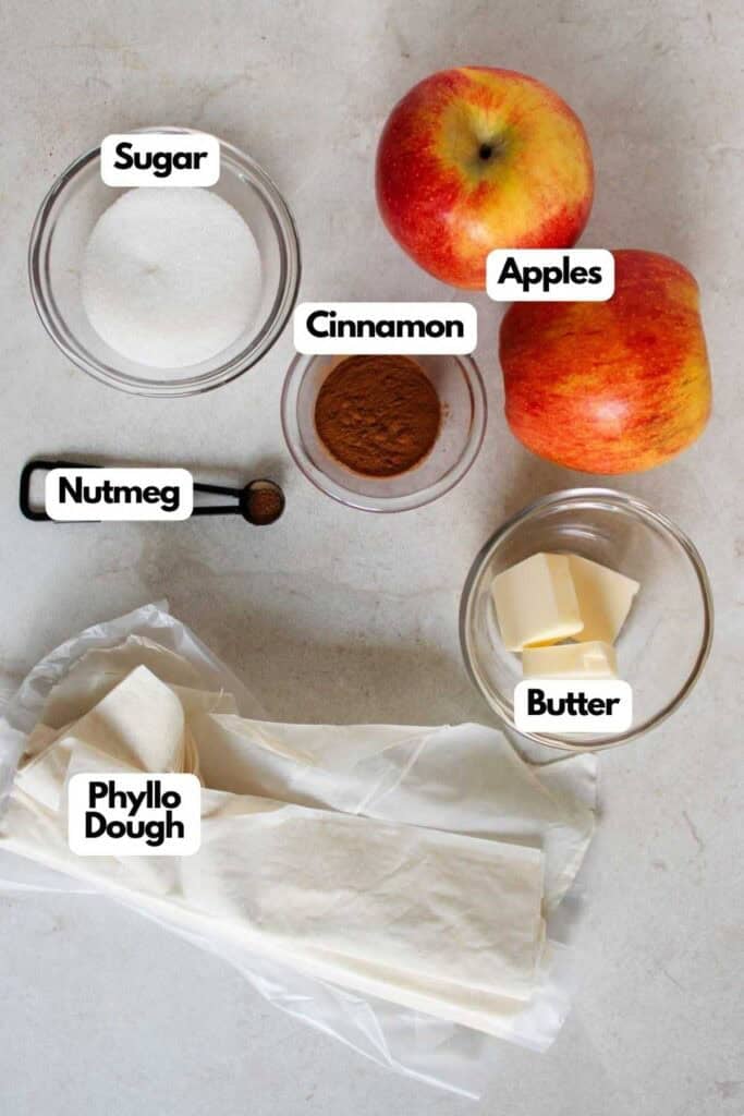 A display of ingredients labeled for making cinnamon apple strudel slices: sugar, apples, cinnamon, nutmeg, butter, and phyllo dough arranged on a countertop.