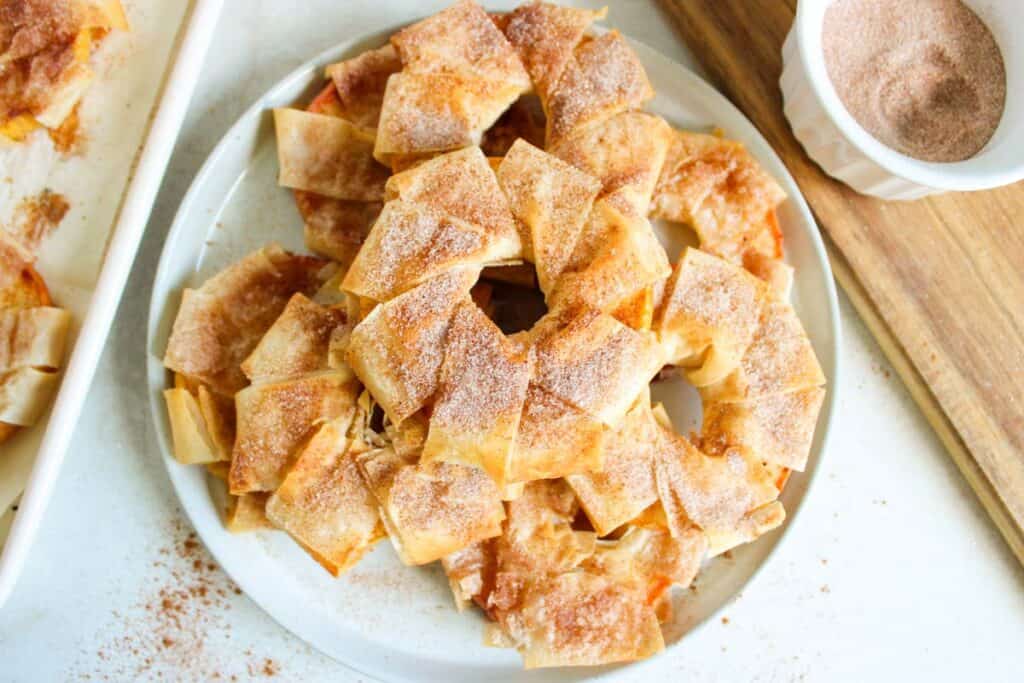 A plate of Cinnamon Apple Strudel slices coated with cinnamon sugar next to a bowl of cinnamon sugar on a wooden board.