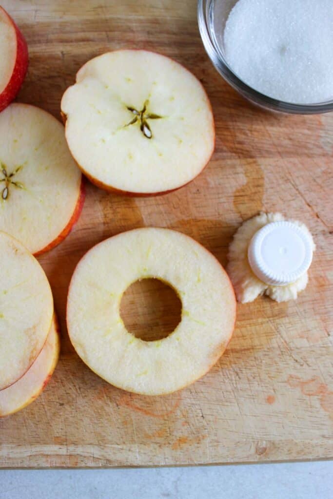 Sliced apples on a cutting board, with one slice cored, evoke the makings of delectable cinnamon apple strudel slices. A bowl of granulated sugar and an apple corer are placed nearby.