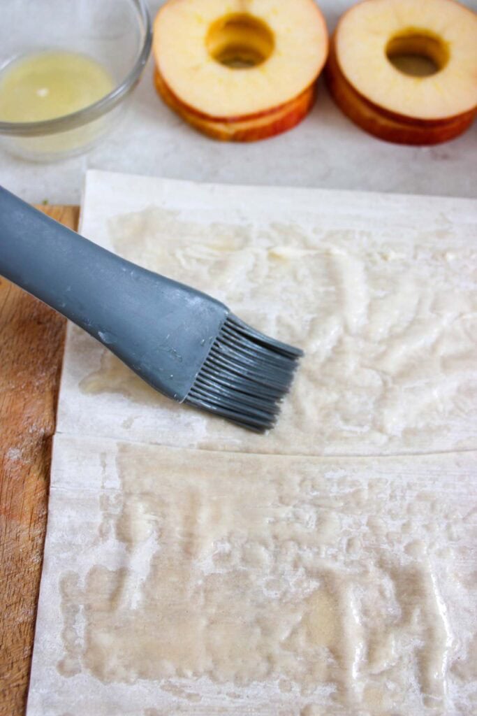 A silicone brush is applied to a pastry sheet with a liquid in a bowl nearby. In the background, sliced apple rings hint at delicious Cinnamon Apple Strudel slices.