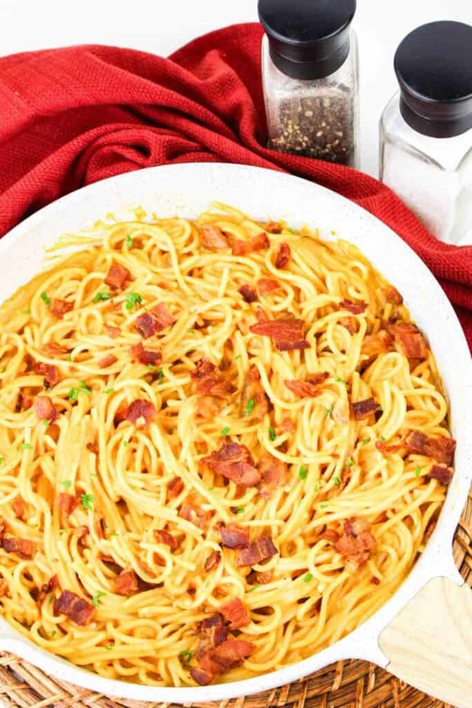 A skillet filled with creamy Pumpkin Carbonara topped with bacon pieces. Two spice shakers and a red cloth are in the background.
