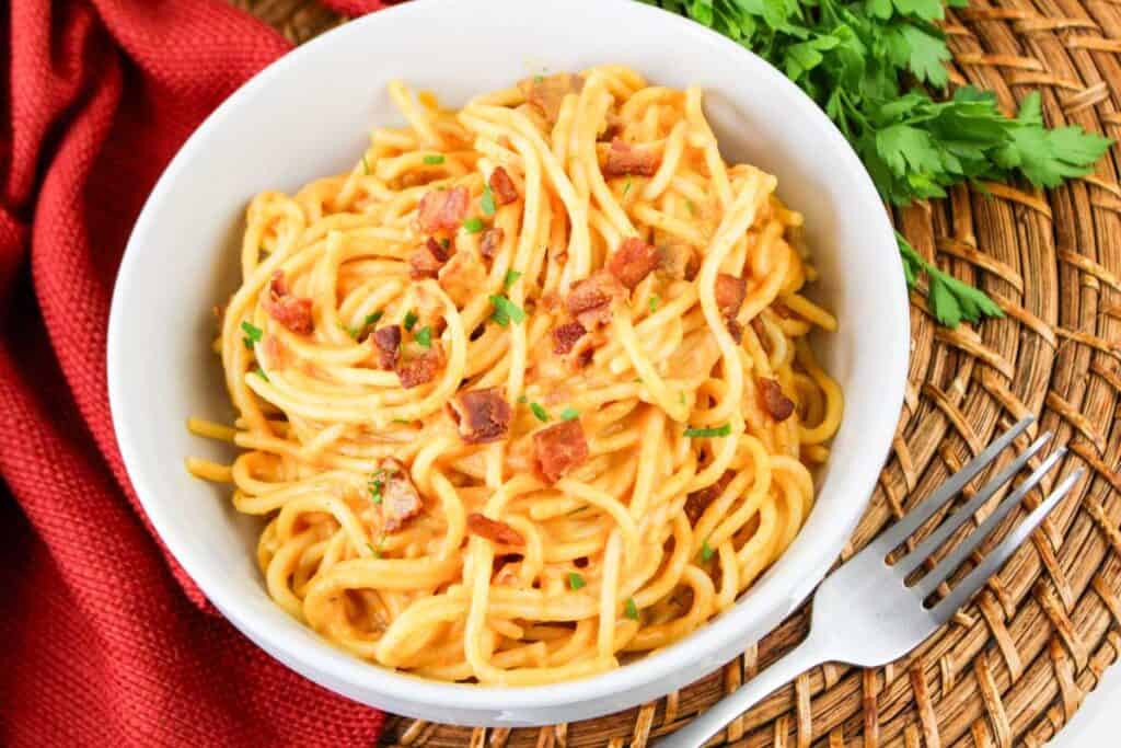 A bowl of Pumpkin Carbonara garnished with chopped herbs and bacon bits placed on a wicker mat, with a red cloth napkin and fork beside it.