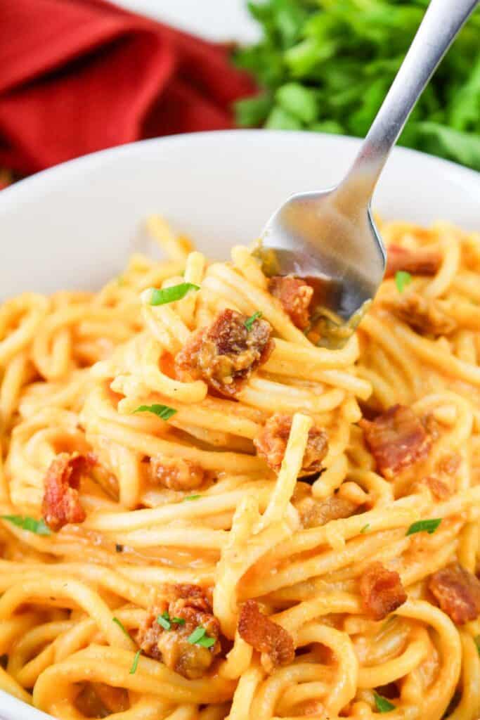 A fork twirls creamy Pumpkin Carbonara with pieces of bacon in a white bowl, placed near a bundle of fresh herbs and a red cloth.