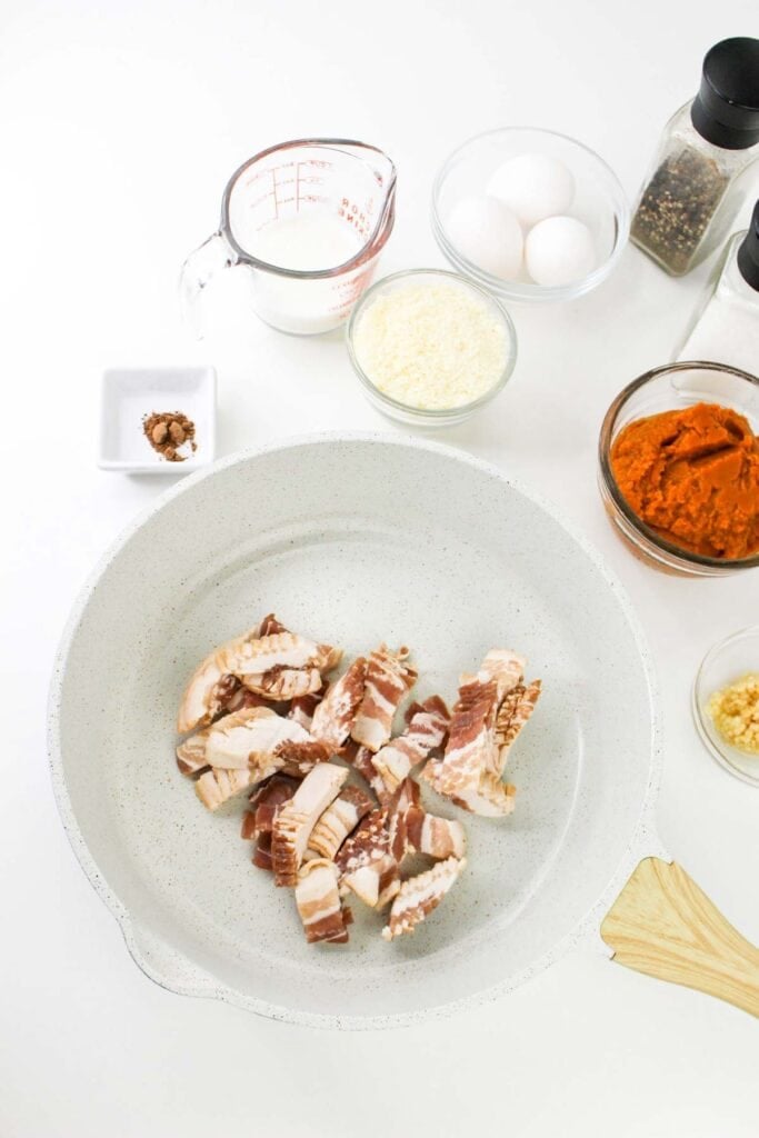 A white pan with diced bacon, surrounded by ingredients for a delicious Pumpkin Carbonara&mdash;including eggs, grated cheese, two glass containers of spices, a measuring cup of milk, a small dish of brown spice, and pumpkin puree.