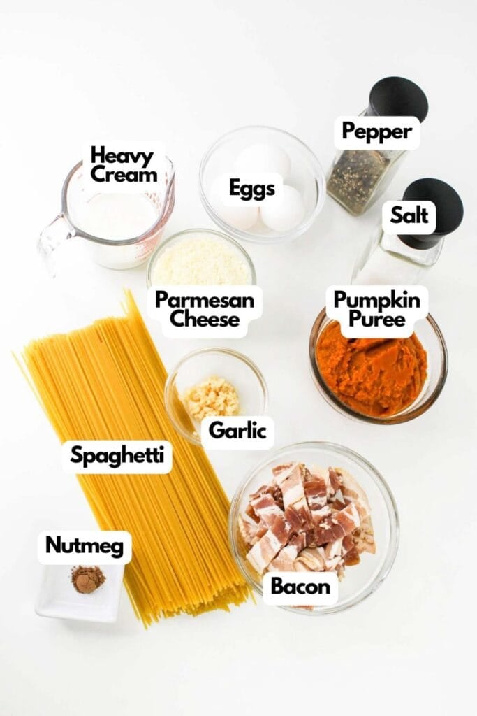 Ingredients for a delightful Pumpkin Carbonara laid out on a white surface include heavy cream, eggs, parmesan cheese, garlic, pepper, salt, pumpkin puree, spaghetti, bacon, and nutmeg.