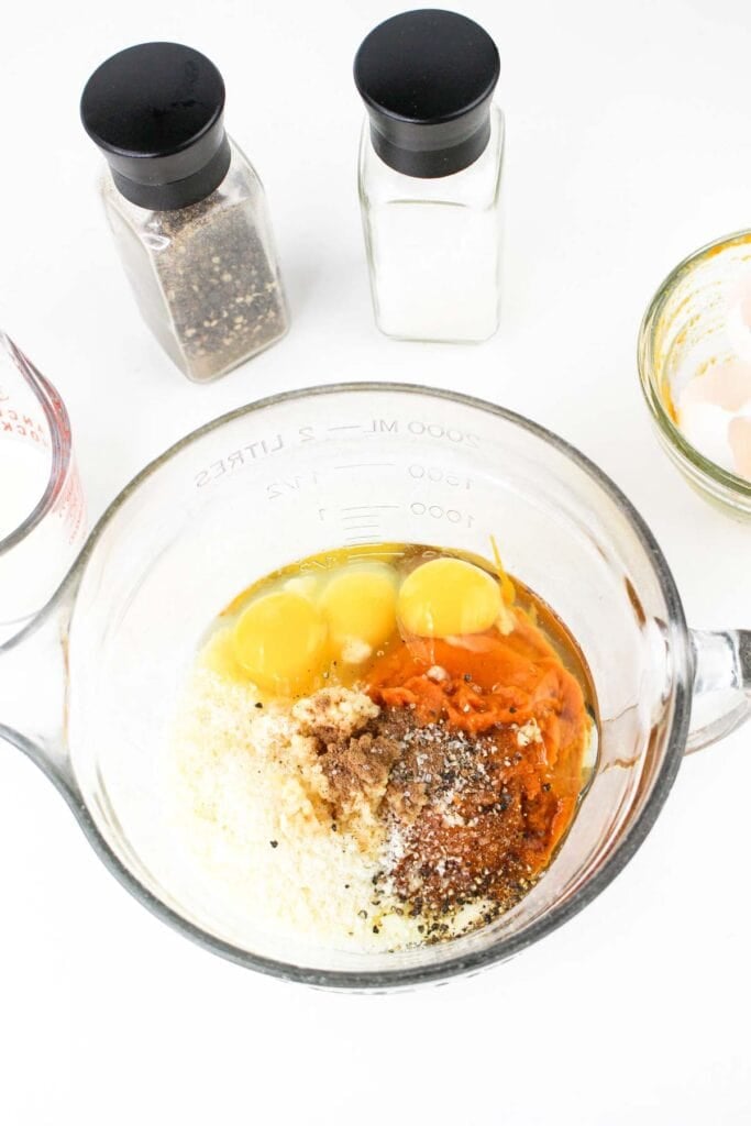 A mixing bowl containing eggs, grated carrots, grated cheese, spices, and seasonings reminiscent of Pumpkin Carbonara. Two spice containers are in the background.