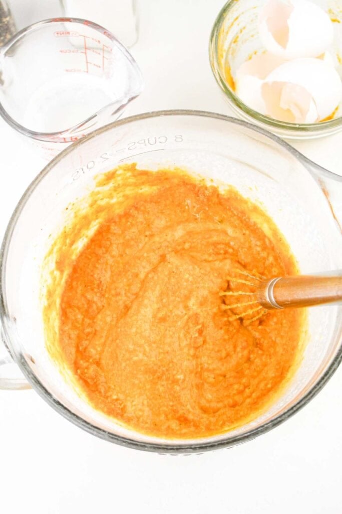 A mixing bowl contains an orange batter being whisked, reminiscent of a rich Pumpkin Carbonara sauce. Behind the bowl, cracked egg shells and a measuring cup with liquid add to the scene's culinary charm.