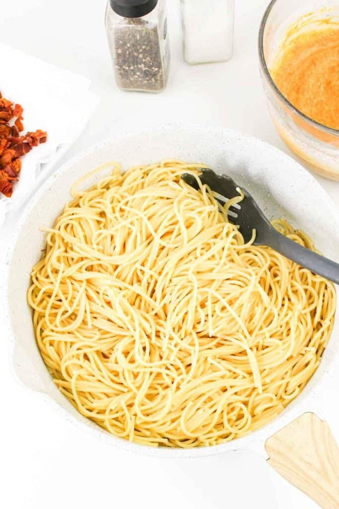 A pot filled with cooked spaghetti, a black pasta spoon, and a bowl of rich pumpkin carbonara sauce beside it. A container of black pepper and another of salt are in the background.