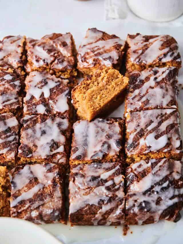 A tray of frosted, square-cut, spiced cake pieces with one piece slightly raised up to reveal its interior.