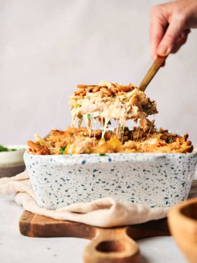 A hand lifting a serving of casserole with melted cheese and a crispy topping from a speckled ceramic dish.