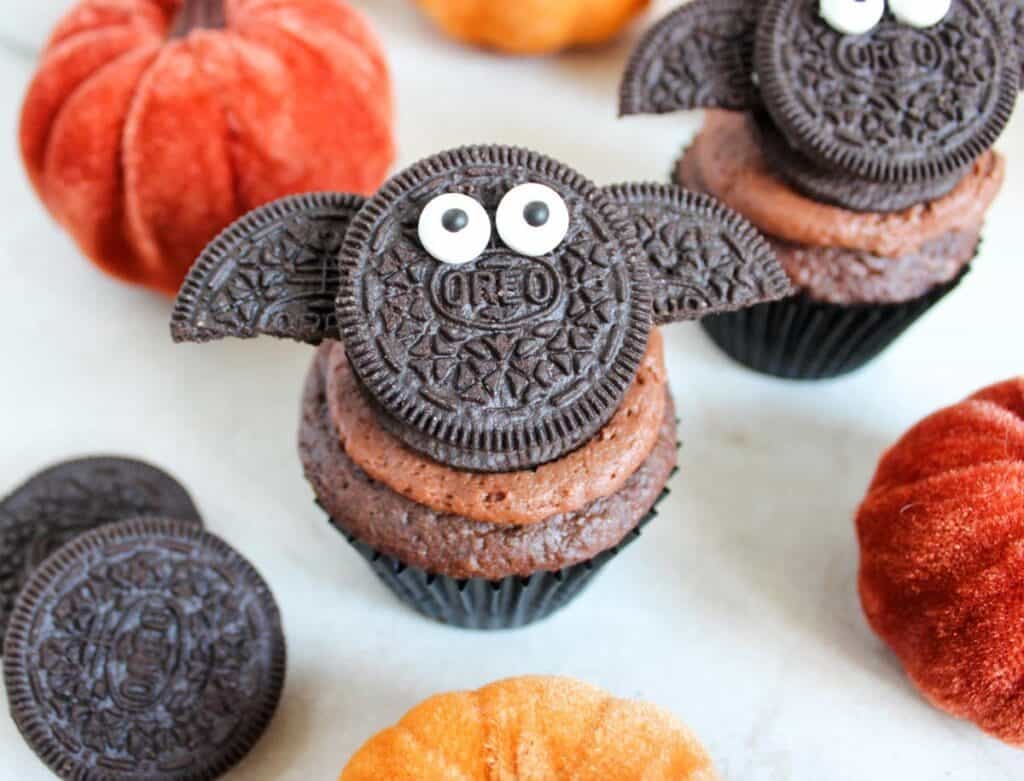 Halloween bat Oreo cupcakes decorated with Oreo cookie wings and candy eyes on a chocolate frosted cupcake surrounded by pumpkin decorations