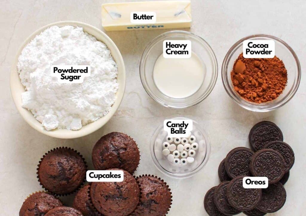 Bat Oreo cupcake ingredients laid out including chocolate cupcakes, Oreo cookies, candy eyes, butter, powdered sugar, cocoa powder, and heavy cream