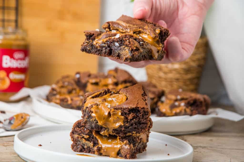 Hand holding a fudgy Biscoff brownie with gooey cookie butter swirl over a stack of brownies on a white plate