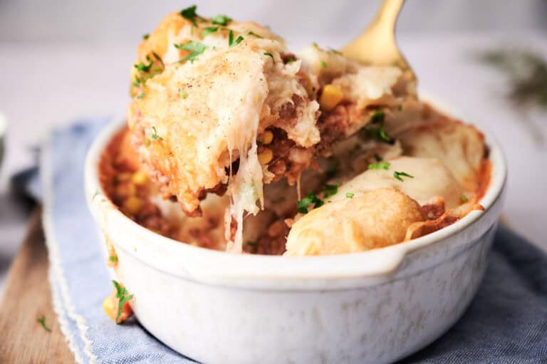 A close-up of a baked Cinnamon French toast casserole dish filled with cheesy, layered pasta or lasagna, topped with fresh herbs, with a spoon lifting a portion.