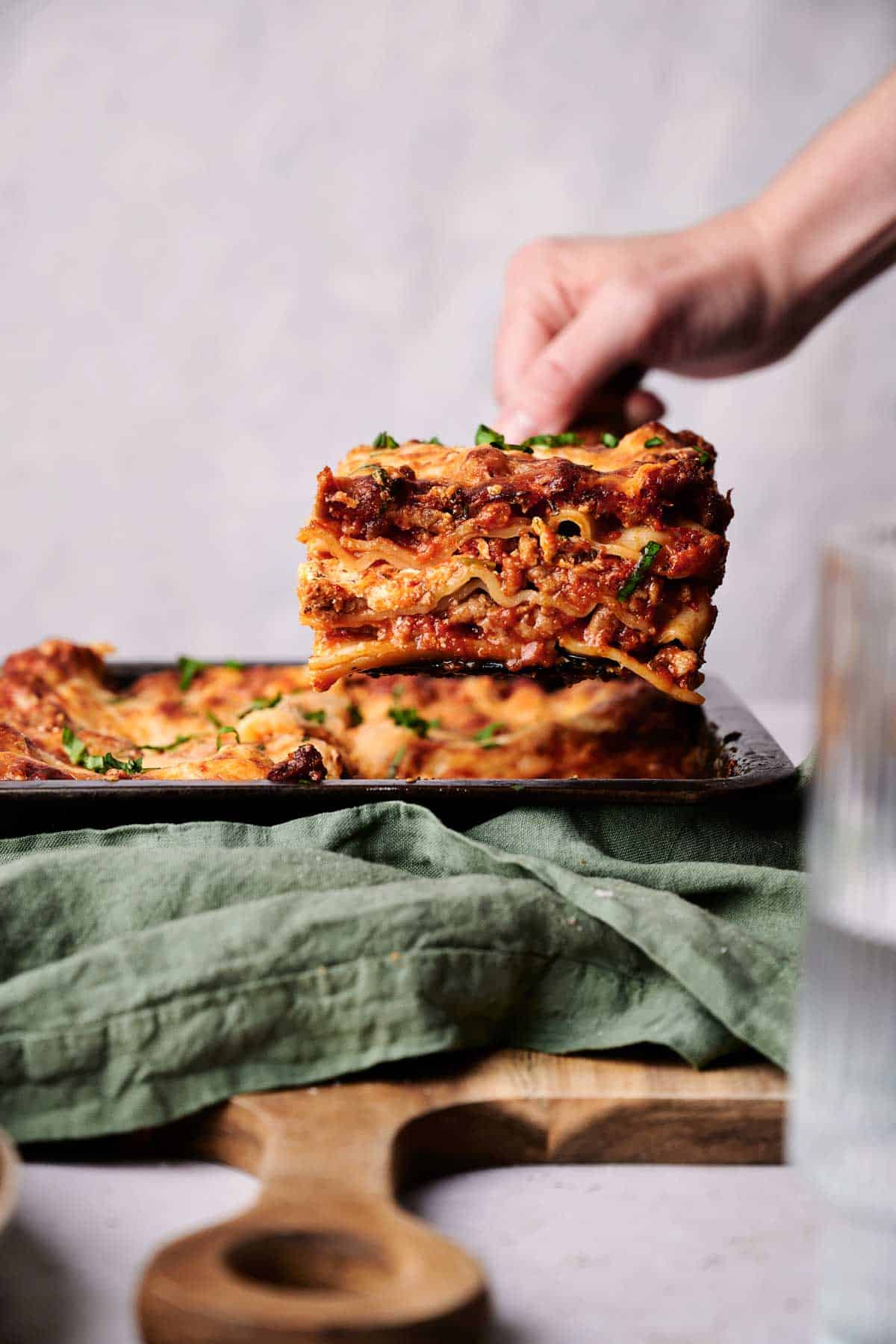 A hand lifting a slice of lasagna from a pan over a green cloth. The lasagna has layers of pasta, meat sauce, and cheese. A wooden cutting board is partially visible in the background.
