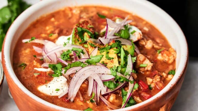 A bowl of chili topped with sliced red onions, avocado, sour cream, and chopped cilantro.