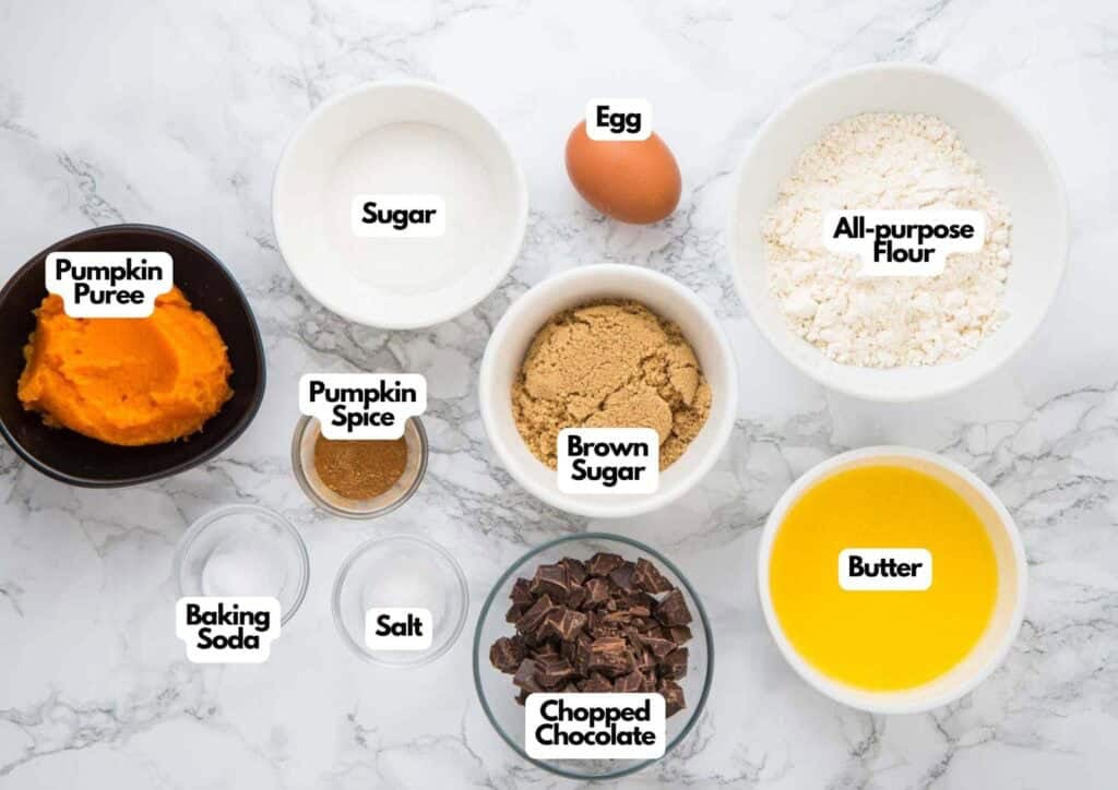 Pumpkin chocolate bar ingredients laid out in bowls including pumpkin puree, chopped chocolate, brown sugar, butter, flour, egg, pumpkin spice, baking soda, and salt