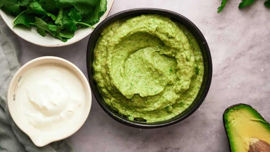 A bowl of guacamole surrounded by cilantro, sour cream, and half an avocado on a marble surface.