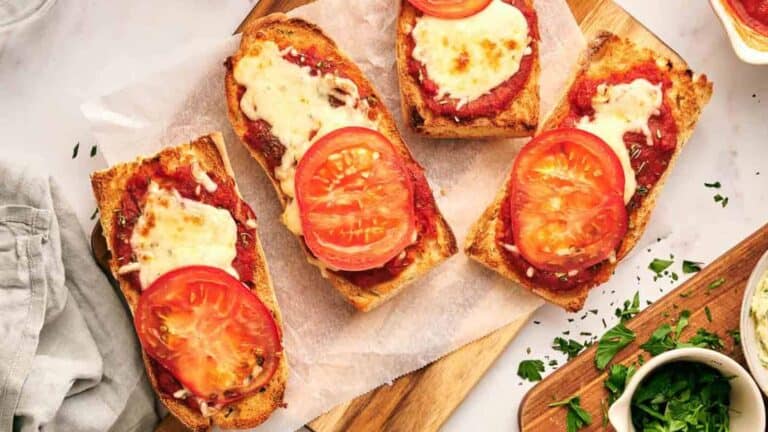 Four toasted baguette slices topped with tomato sauce, melted cheese, and tomato slices on a parchment-lined wooden board.