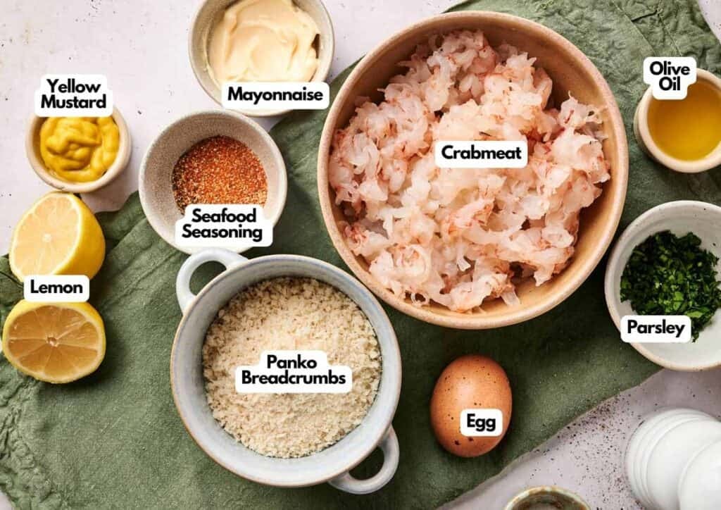 Flat lay of Maryland crab cake ingredients including crabmeat, panko breadcrumbs, egg, mayonnaise, yellow mustard, lemon, parsley, seafood seasoning, and olive oil
