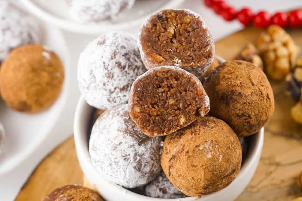 Homemade rum balls coated in powdered sugar and cocoa powder, piled in a white bowl, with one cut open to show the chocolate walnut interior