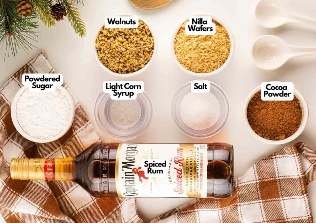 Rum ball ingredients laid out on a white surface including walnuts, Nilla wafers, powdered sugar, light corn syrup, salt, cocoa powder, and Captain Morgan Spiced Rum