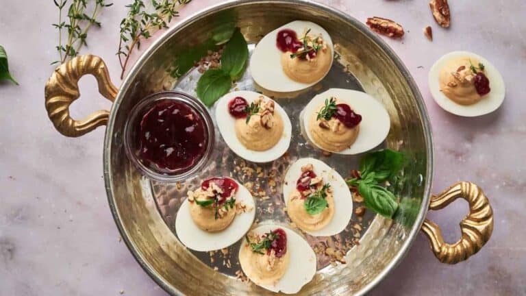 Deviled eggs on a platter, topped with herbs and a red sauce, with a small dish of sauce on the side. Basil leaves and pecans are scattered around.