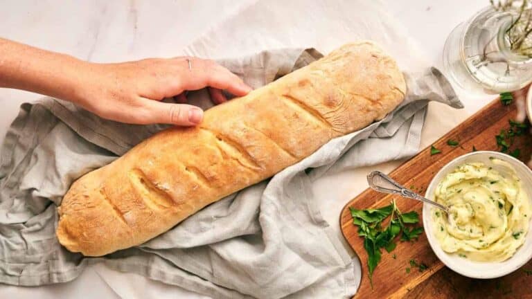 A hand reaches for a freshly baked baguette on a cloth. Nearby, a cutting board holds a bowl of herb butter and parsley garnishes.