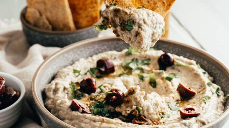 A close-up of baba ganoush in a bowl, topped with herbs and olives. A pita chip is dipped into the creamy texture.