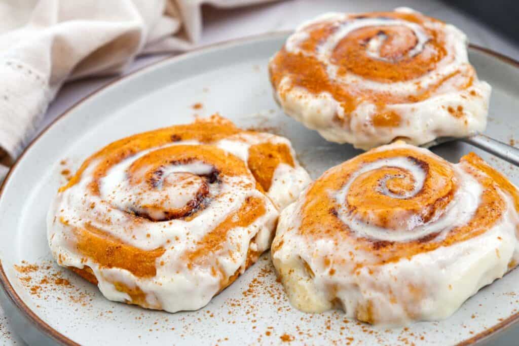 Three pumpkin cinnamon rolls smothered in cream cheese frosting and dusted with cinnamon, served on a gray plate