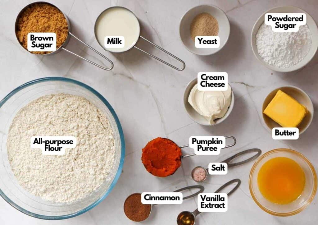 Pumpkin cinnamon roll ingredients laid out on a marble surface including all-purpose flour, pumpkin puree, cream cheese, butter, milk, brown sugar, yeast, powdered sugar, cinnamon, vanilla extract, and salt