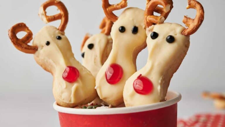Festive cookies decorated as reindeer with pretzel antlers and candy noses in a red cup.
