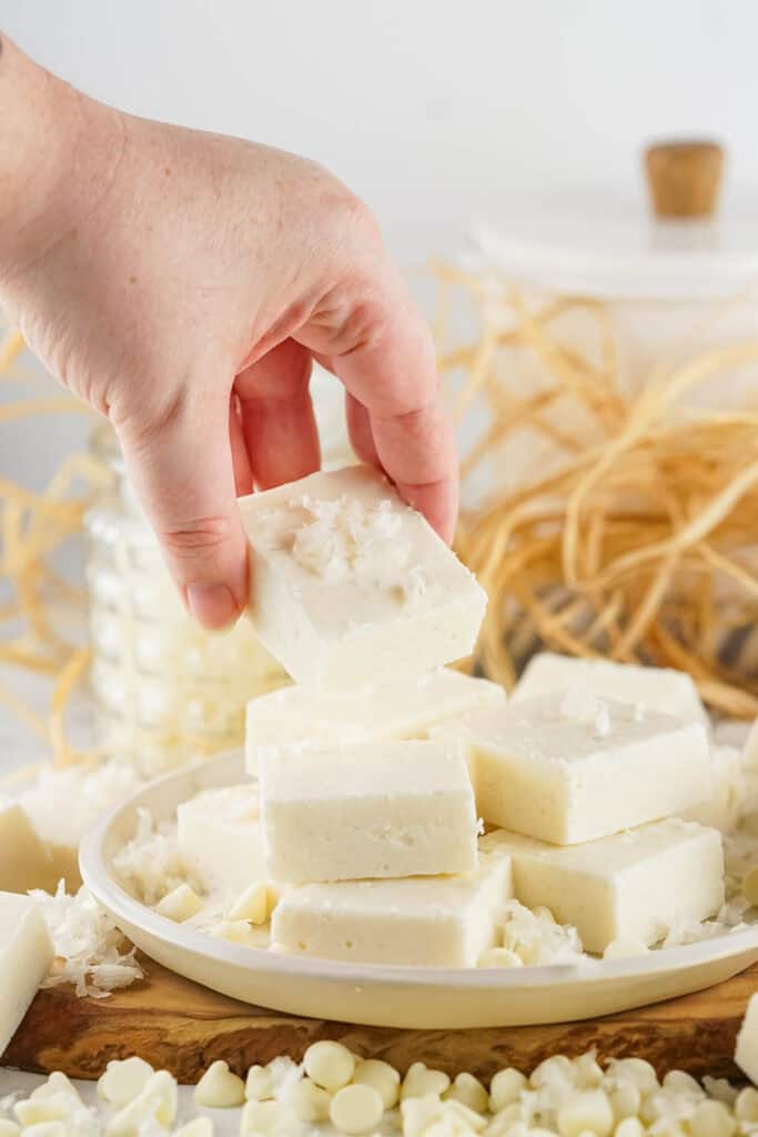 A hand picks up a square piece of coconut fudge from a plate stacked with similar pieces, surrounded by white chocolate chips and shredded coconut.