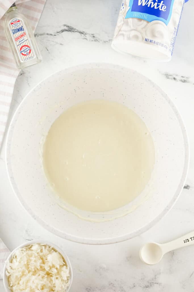 A bowl of white frosting sits on a marble surface, accompanied by a container of rich coconut fudge, coconut flakes in a bowl, a teaspoon, and a bottle of peppermint extract.