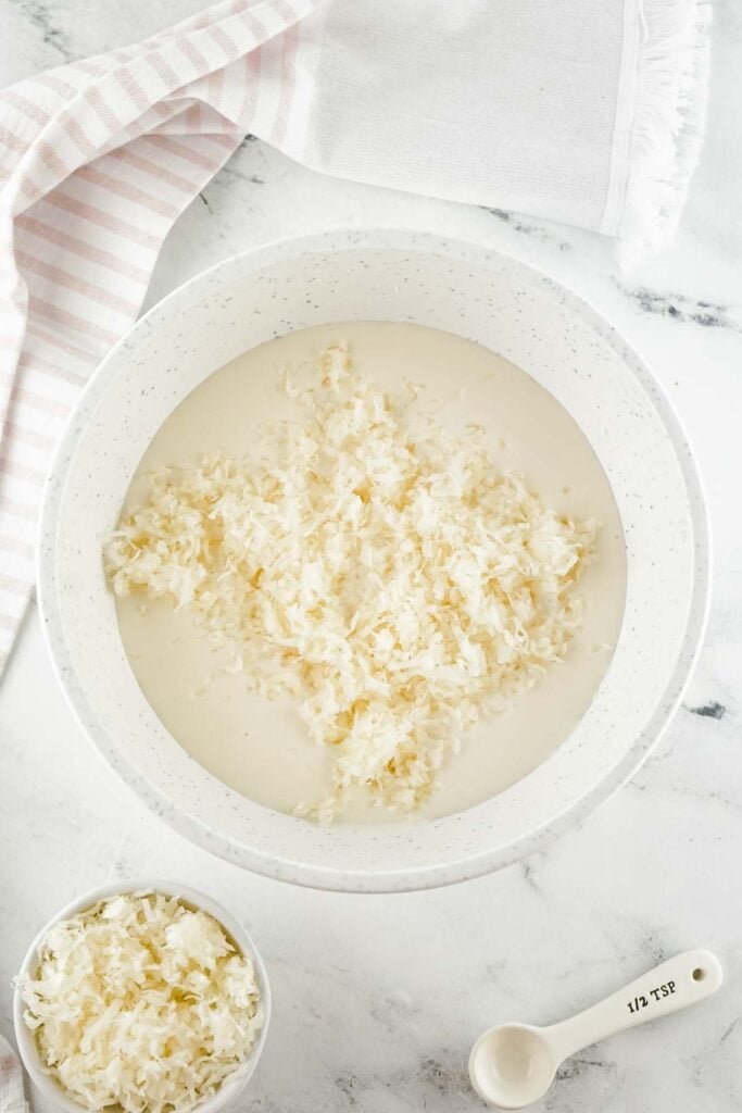A white bowl with soaking shredded dried fungus rests on a marble surface, accompanied by a small bowl of fungus and a 1/2 teaspoon measuring spoon, hinting at an exotic recipe perhaps enriched with creamy coconut fudge.