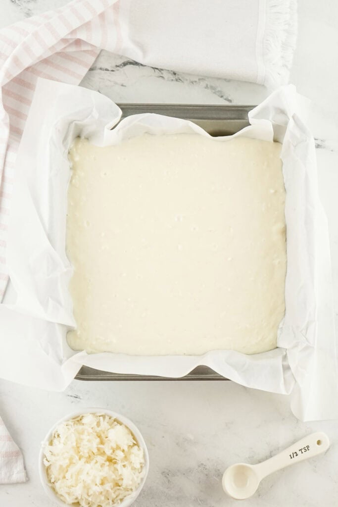 A square baking dish lined with parchment paper cradles a luscious white batter, poised to transform into delectable coconut fudge. Nearby, a bowl of shredded coconut and a measuring spoon stand ready to elevate this sweet creation.
