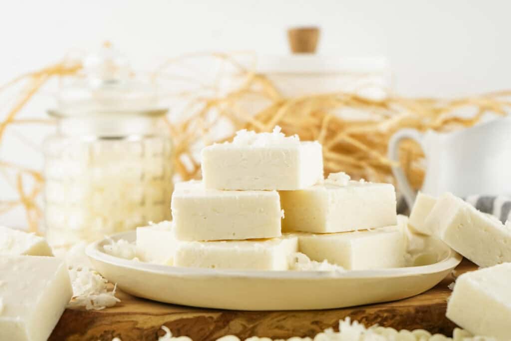 A plate of stacked white soap bars, reminiscent of creamy coconut fudge, rests on a wooden surface, with jars and dried decorative elements softly framing the background.
