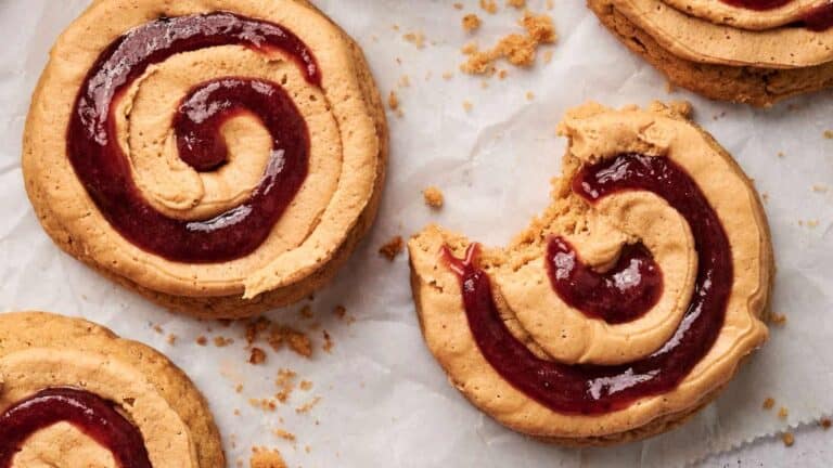 Cookies with spiral patterns of red jam on a light brown surface, with one cookie partially eaten and crumbs scattered around.