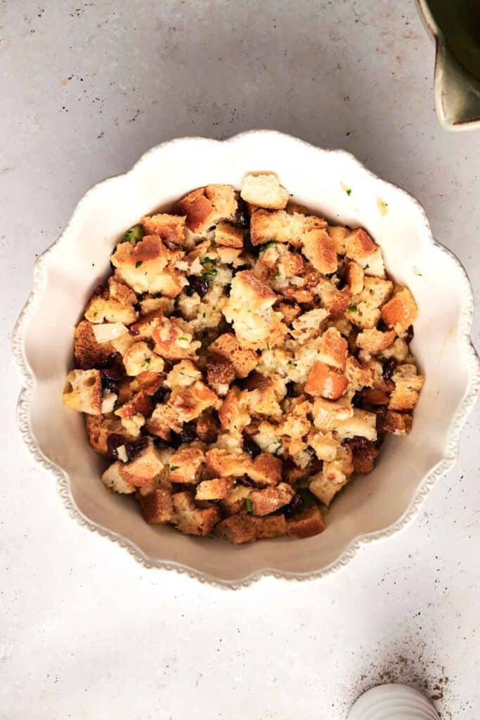 A white bowl brimming with delicious bread stuffing, featuring seasoned, cubed bread mixed with aromatic herbs, rests on a light surface.