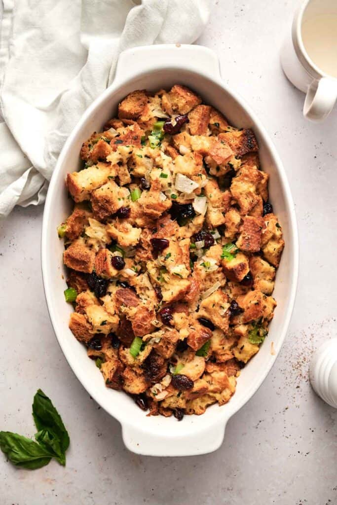 A delightful dish of baked bread stuffing, featuring celery and cranberries, rests in an oval vessel on a light surface, accompanied by a white napkin and adjacent cup.