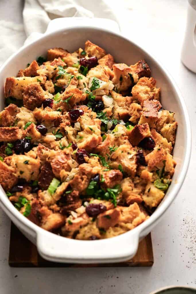 A casserole dish brimming with savory bread stuffing, showcasing chunks of toasted bread, cranberries, chopped herbs, onions, and celery.