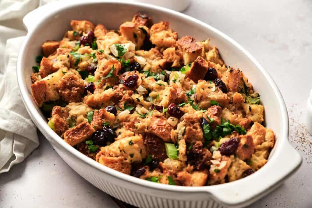 A white oval dish brimming with seasoned bread stuffing, artfully mixed with herbs, celery, and tart cranberries.