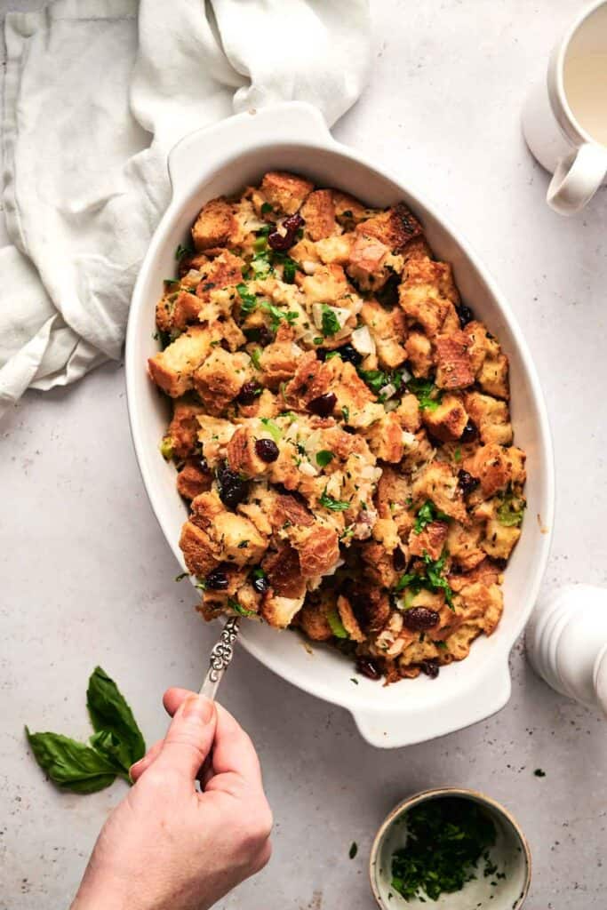 A hand elegantly serving fragrant bread stuffing from a pristine white oval dish on a light surface, perfectly complemented by a crisp white napkin and a small bowl of fresh herbs.