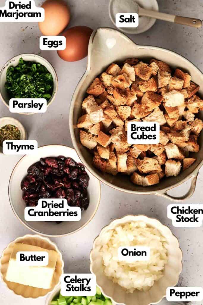 Ingredients for a delicious bread stuffing recipe laid out on a surface: eggs, salt, bread cubes, dried marjoram, parsley, thyme, chicken stock, dried cranberries, butter, celery stalks, onion, and pepper.