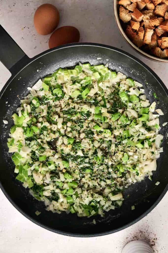Chopped green peppers and onions are saut&eacute;ing in a black skillet, sprinkled with herbs. Two whole eggs sit beside a bowl of croutons, ready to be transformed into a savory bread stuffing.