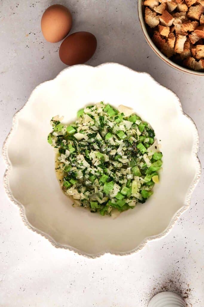 Mixed chopped vegetables in a white scalloped bowl, accompanied by two eggs and a bowl of croutons for homemade bread stuffing, all arranged on a light surface.
